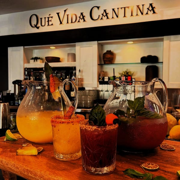 Two pitchers and two glasses of colorful Mexican drinks on a wooden bar under a "Qué Vida Cantina" sign.