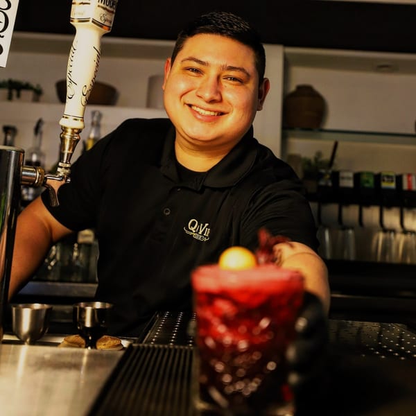 Smiling Que Vida Tacos bartender serving a vibrant red cocktail with a lemon garnish.