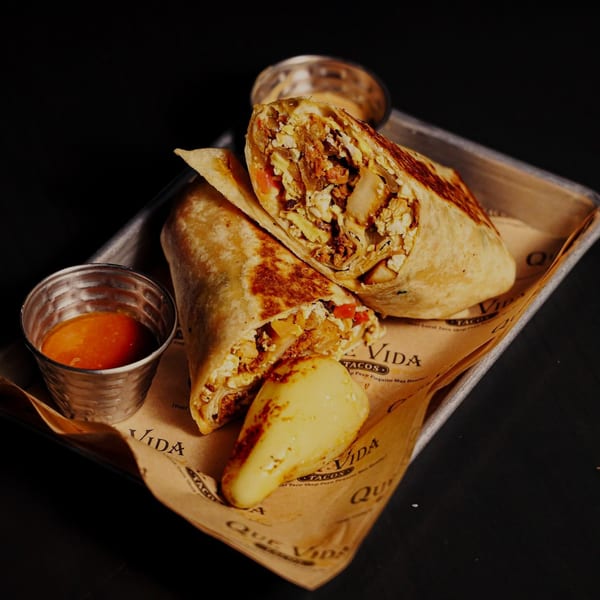 Two Que Vida Tacos burritos, one cut open, with a side of sauce on a metal tray.