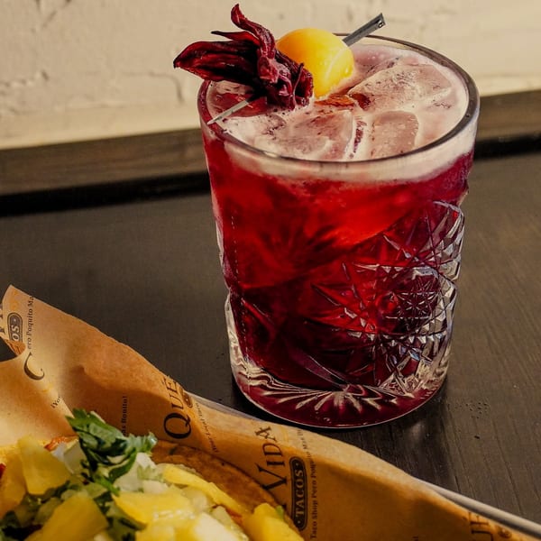 A Que Vida Tacos taco in branded paper next to a red cocktail with a lemon and hibiscus garnish.