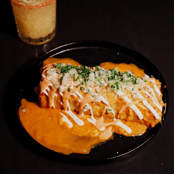 A Que Vida Tacos burrito covered in orange sauce, white crema, diced onions, and cilantro, with a drink.