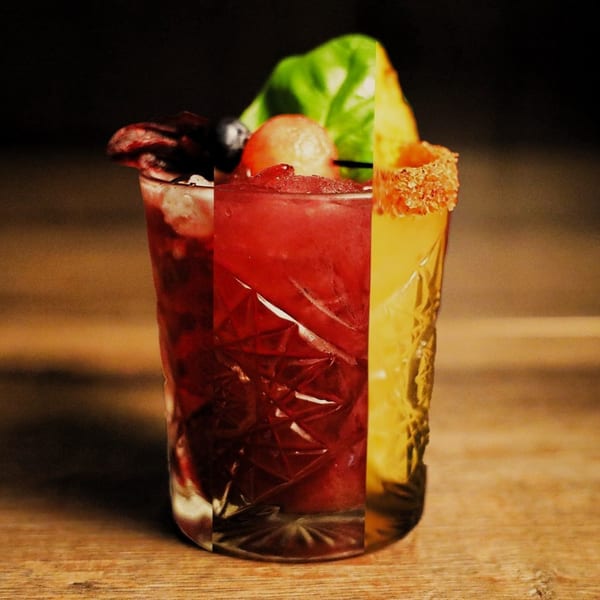 A vibrant red and yellow cocktail in a textured glass, garnished with fruit and basil on a wooden table.