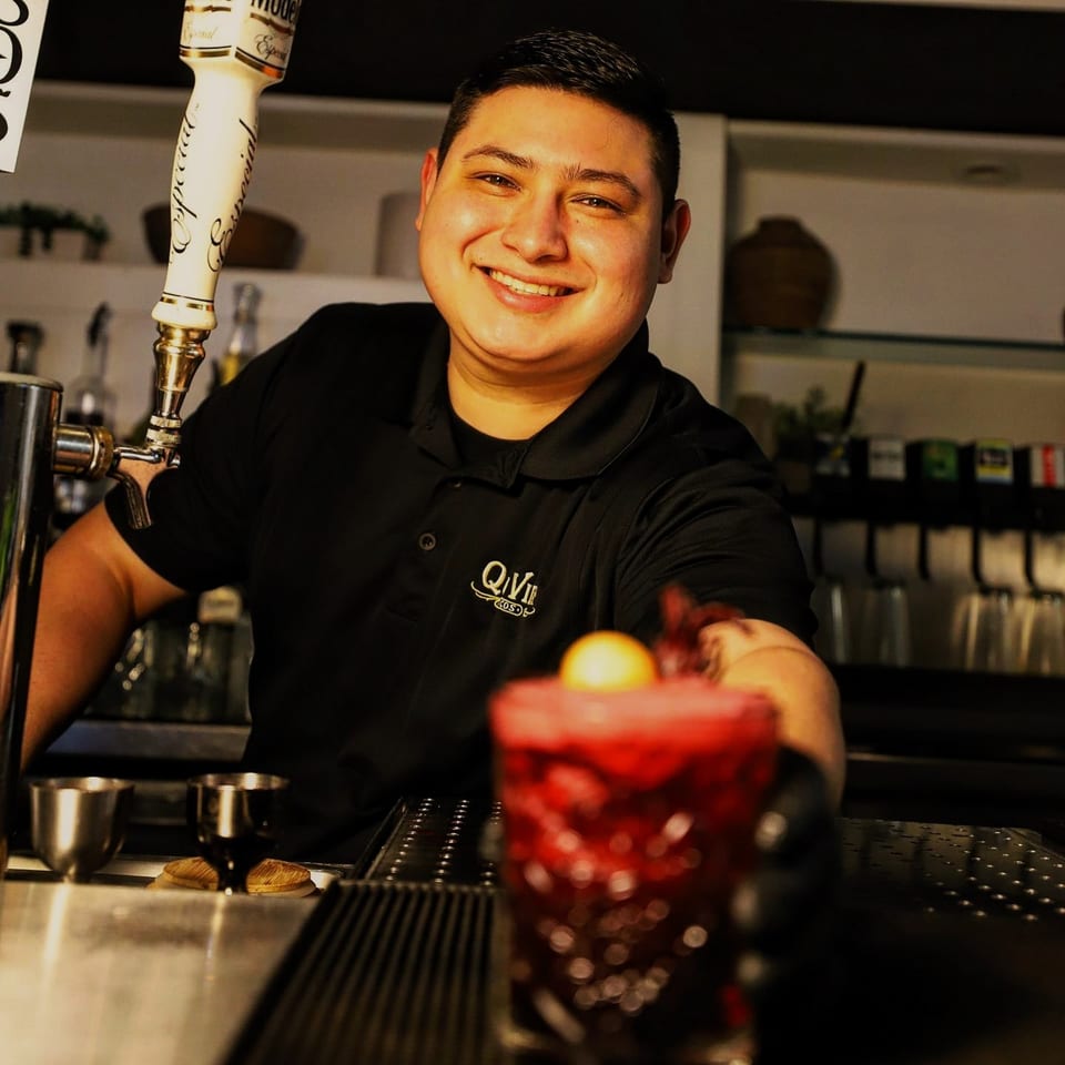 Smiling Que Vida Tacos bartender serving a vibrant red cocktail with a lemon garnish.